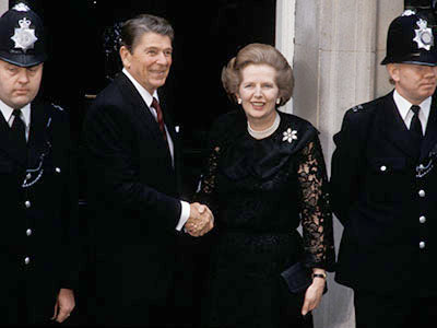 Two individuals shake hands while flanked by two uniformed officers. The person on the left is in a suit, and the person on the right wears a formal dress. Both are smiling as they stand in front of a building entrance.